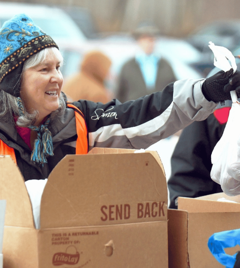 Outside in the winter, a volunteer woman at a mobile food pantry is handing a bag of food to a neighbor