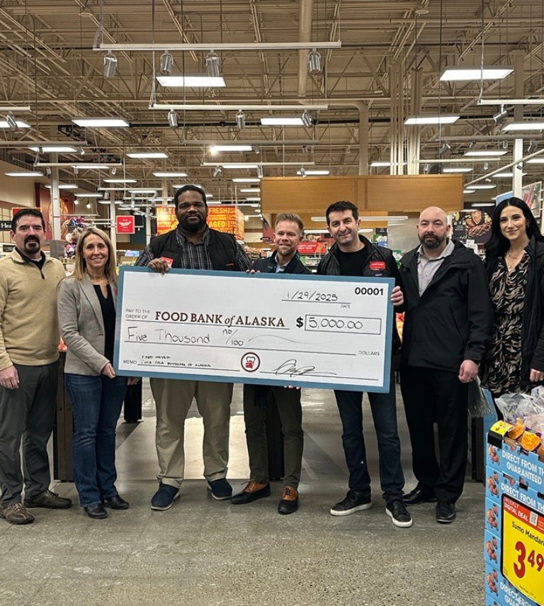 A group of 5 men and two woman standing in a grocery store with a check to Food Bank of Alaska for $5,000